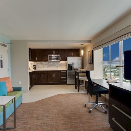 A modern living area with a kitchenette, sofa, TV, desk, and a large window showing a city view. Warm, inviting atmosphere.