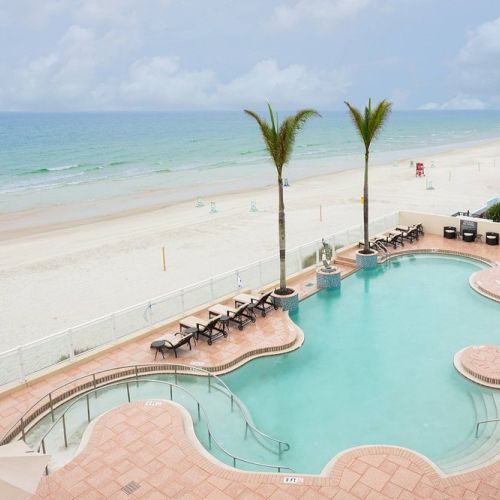 A beachside view with a curved pool, loungers, two palm trees, and a sandy beach leading to the ocean under cloudy skies.