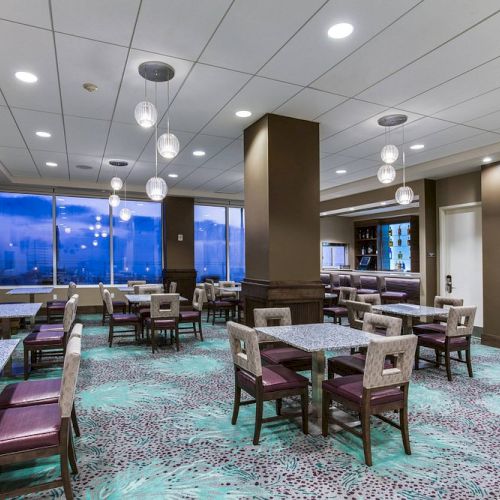 A modern dining area with tables, chairs, pendant lights, large windows, and a bar in the corner. The space appears clean and inviting.