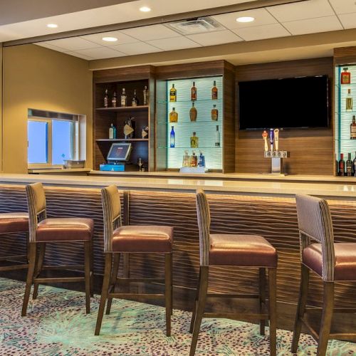 This image shows a modern bar setting with a counter, tall chairs, assorted bottles on shelves, and a TV on the wall.