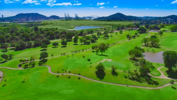 Aerial view of a lush green golf course with sand traps and ponds, surrounded by hills and industrial cranes in the distance.