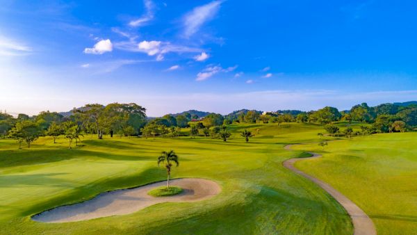 A lush green golf course with sand bunkers, winding paths, palm trees, and distant hills under a blue sky.