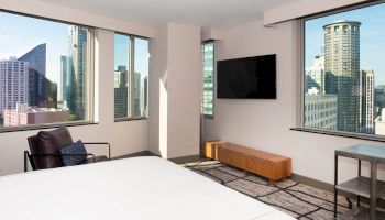A modern hotel room with a large bed, armchair, TV, and stunning views of a city skyline through multiple windows.