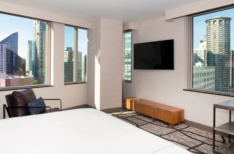 A modern hotel room with a large bed, armchair, TV, and stunning views of a city skyline through multiple windows.
