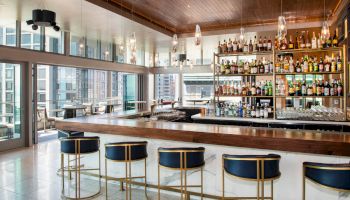 A modern bar with high chairs, a well-stocked drink shelf, hanging lights, and large windows showing a cityscape view.