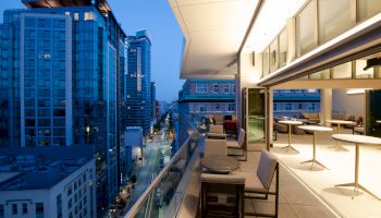 The image shows a modern cityscape at dusk, featuring a high-rise building with a well-lit balcony and outdoor seating area overlooking the street.