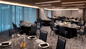 A banquet hall is set up with round tables and black chairs, arranged for a formal event. Table settings include plates, glasses, and folded napkins.