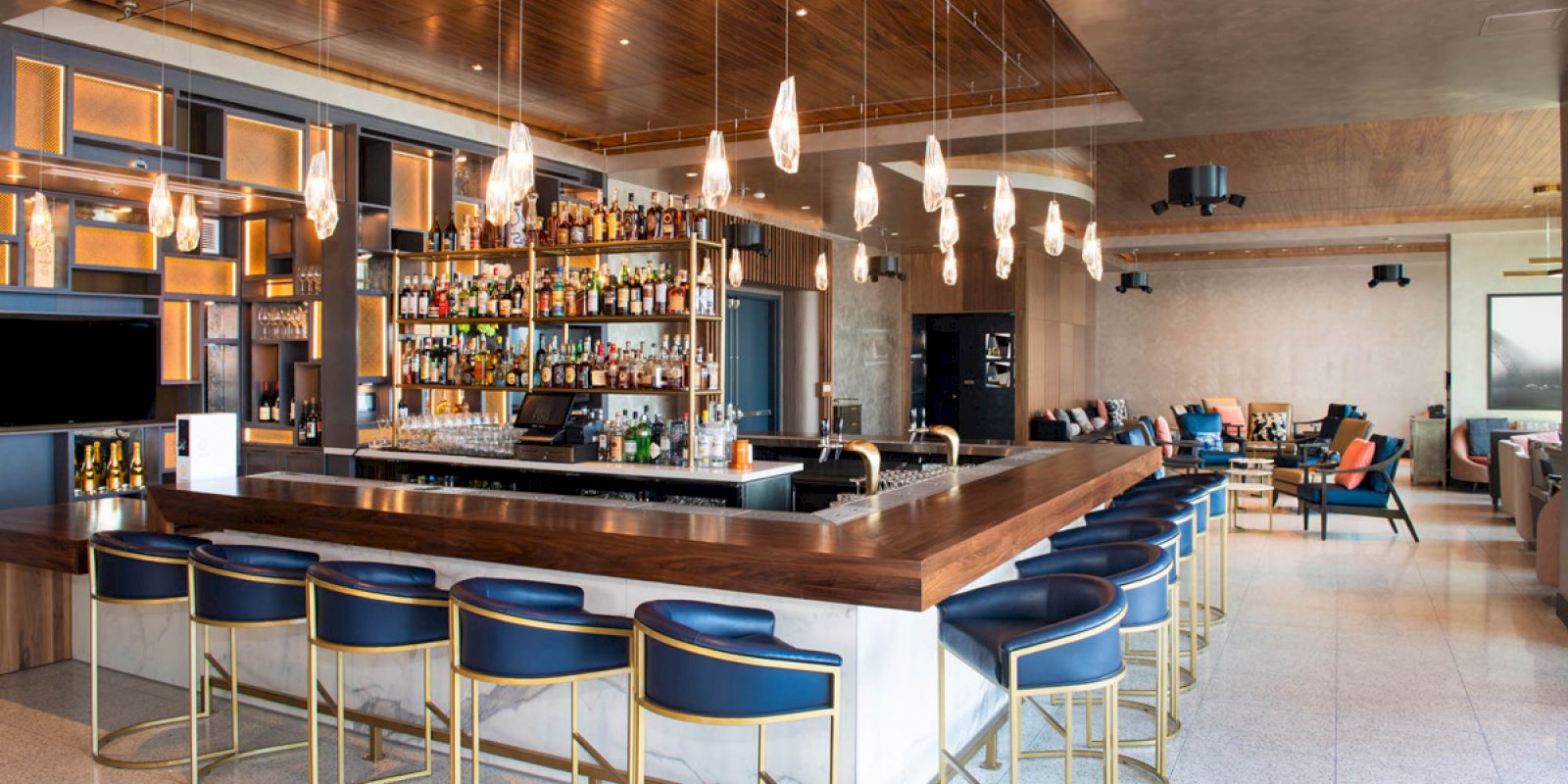 A modern bar with a large counter, blue barstools, hanging lights, and a fully stocked shelf of liquor bottles, set in a stylish lounge area.