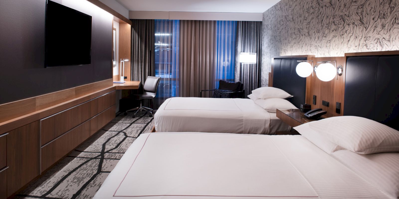 The image shows a modern hotel room with two beds, a large TV, a work desk and chair, and ambient lighting.