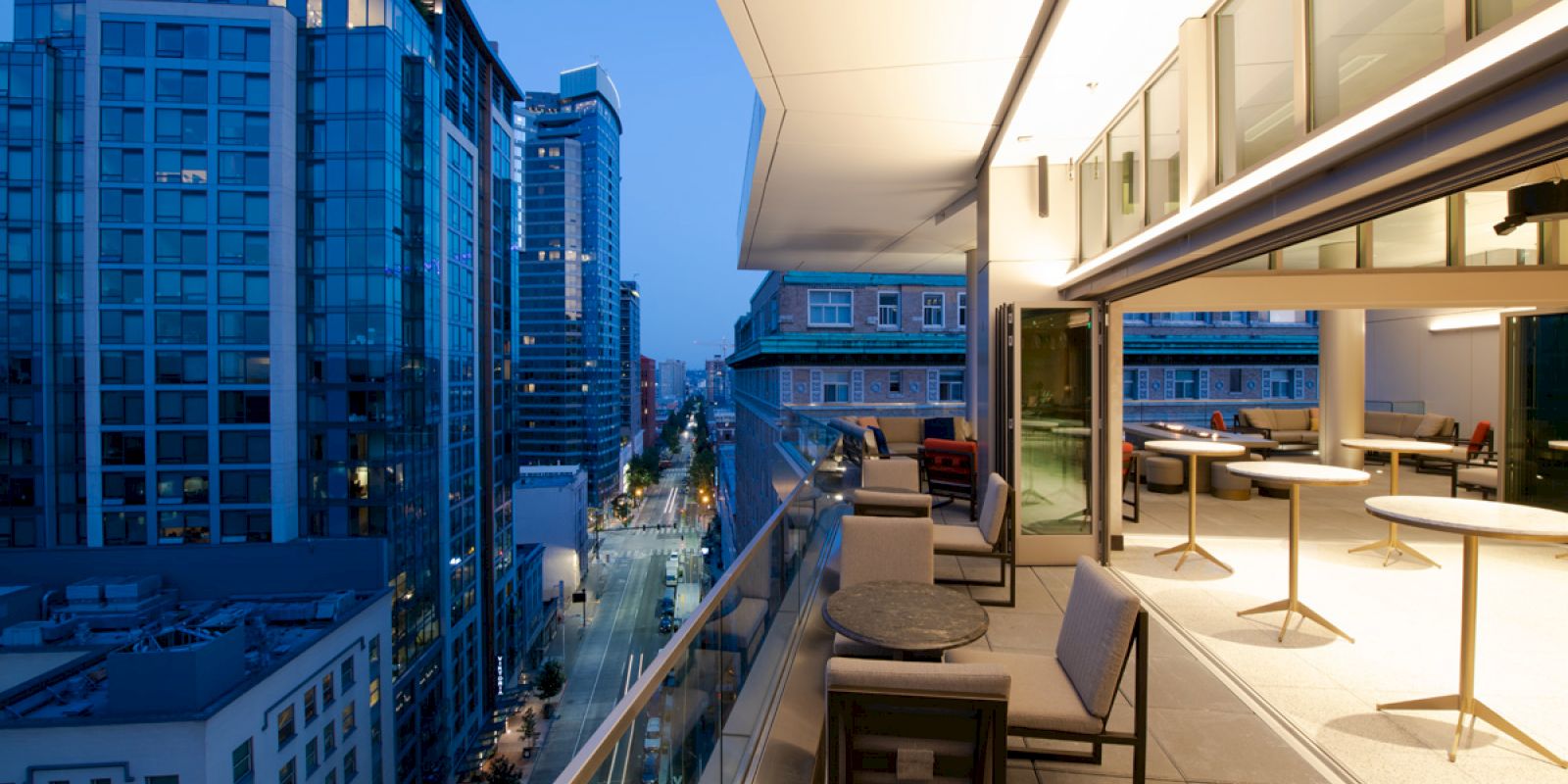 The image shows a modern balcony at dusk with seating and tables, overlooking a city street and surrounded by tall buildings.