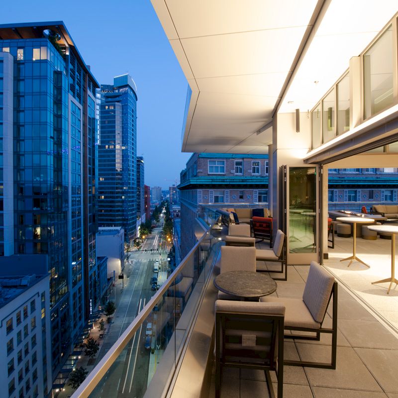 The image shows a modern outdoor balcony area with seating, overlooking a city street and surrounding high-rise buildings during twilight.