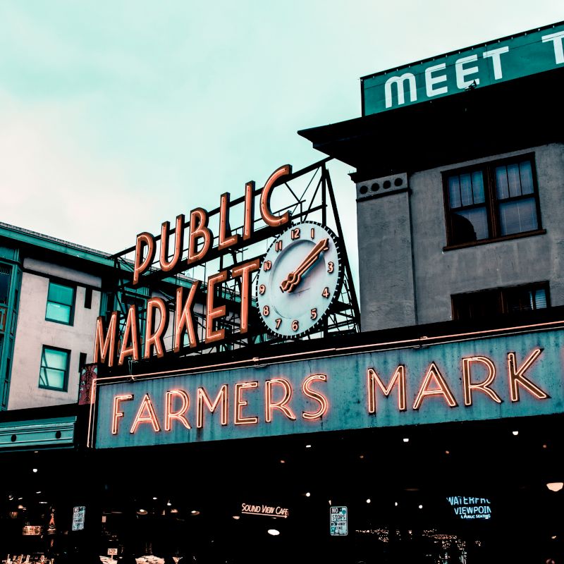 The image shows the neon signs of "Public Market" and "Farmers Market" on a building, likely an entrance to a market area.