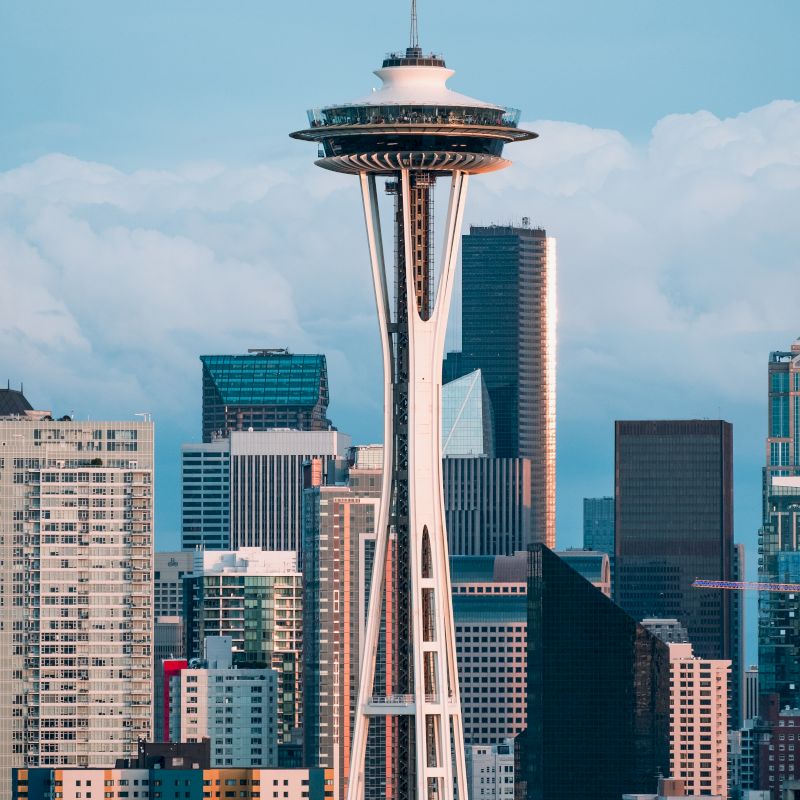The image shows the Seattle Space Needle with a backdrop of the city’s skyline, including various tall buildings and skyscrapers, under a partly cloudy sky.