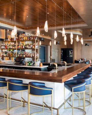 The image shows a stylish bar with modern decor, featuring a central bar counter, blue bar stools, hanging lights, and a well-stocked liquor shelf.