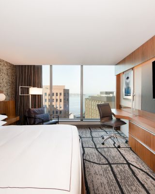 A modern hotel room features a large bed, mounted TV, desk, chair, and large window with a city view, showcasing a contemporary design.