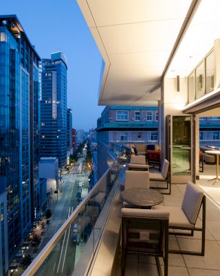 A modern cityscape viewed from a balcony at dusk, with buildings, a lit interior space with seating, and a glass railing along the edge.