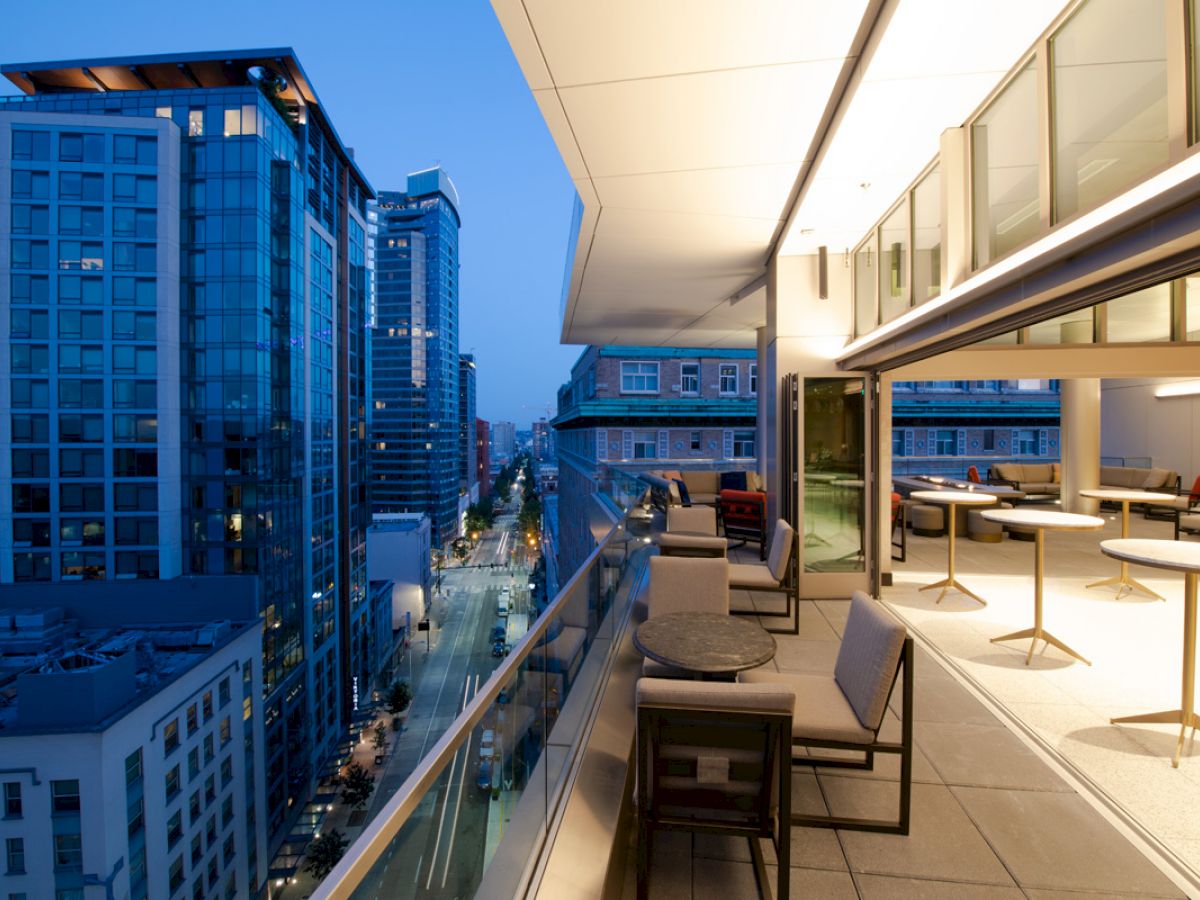 The image shows a modern cityscape at dusk, featuring tall buildings and a stylish balcony with tables and seating overlooking the street below.
