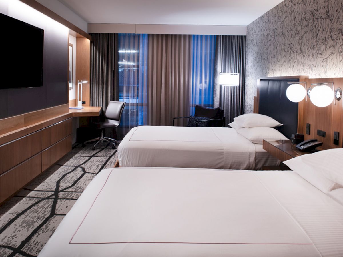 A modern hotel room with two beds, a flat-screen TV, a work desk, a chair, and stylish decor.