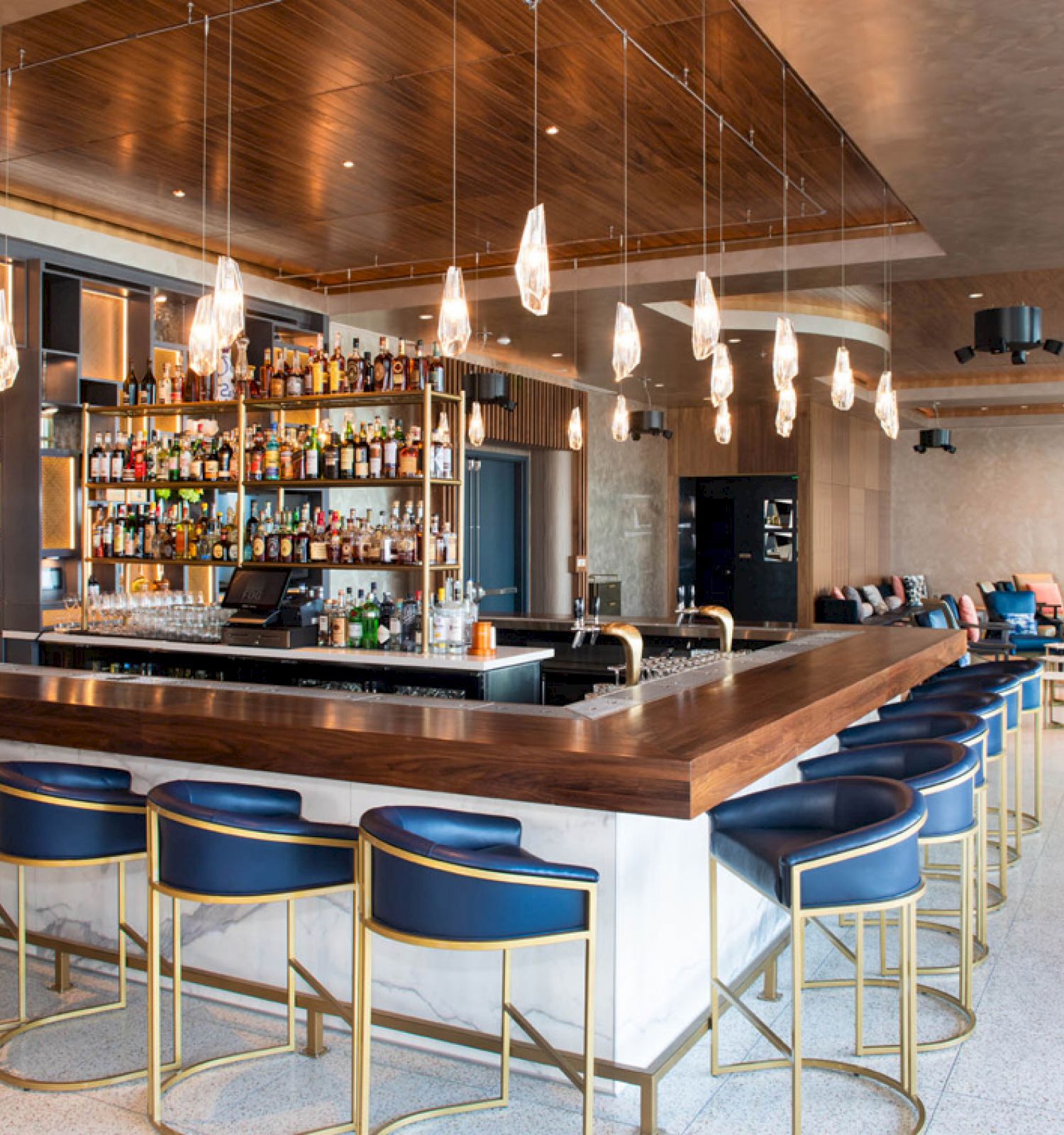 The image shows a modern bar with a large counter, blue bar stools, hanging lights, and a fully stocked liquor shelf in a stylish, well-lit setting.