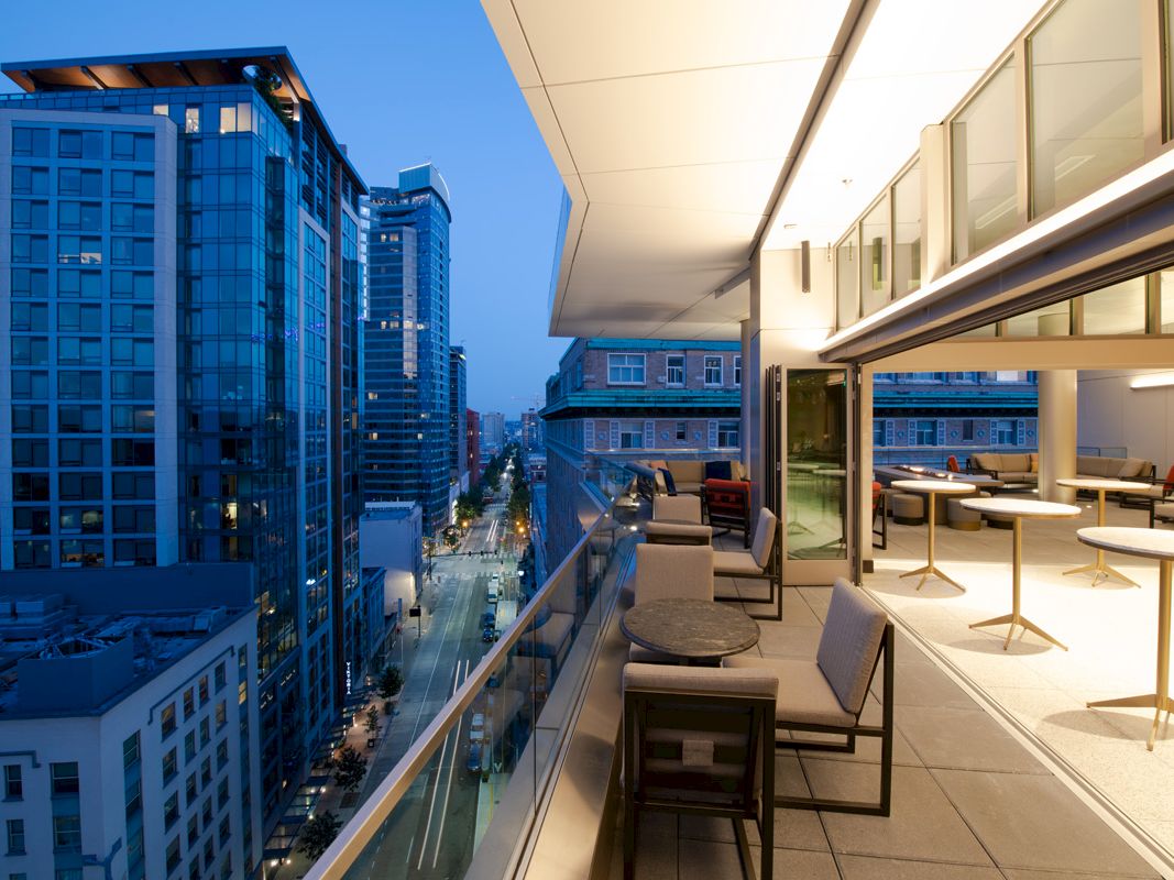 An urban balcony scene at dusk, overlooking a city street and modern buildings, with tables and cushioned seating arranged on the patio.