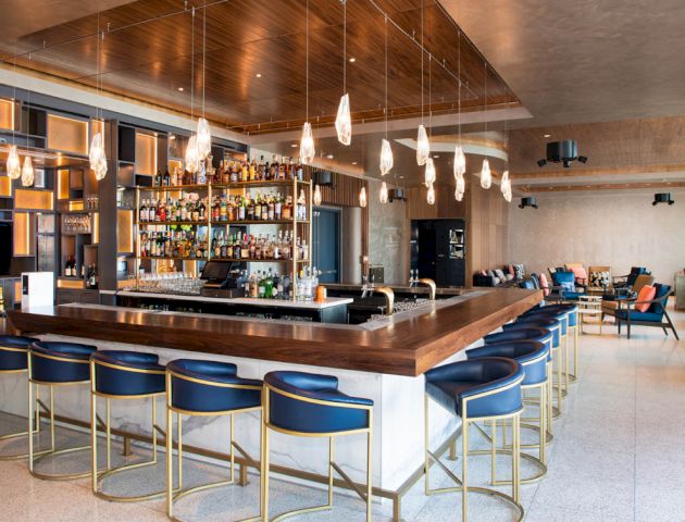 The image shows a modern bar with a wooden countertop, blue chairs, hanging lights, and a stocked liquor shelf. There's a lounge area in the background.