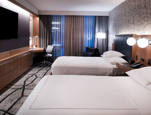 The image shows a modern hotel room with two neatly made double beds, a wall-mounted TV, a study desk with a lamp, and large windows with curtains.