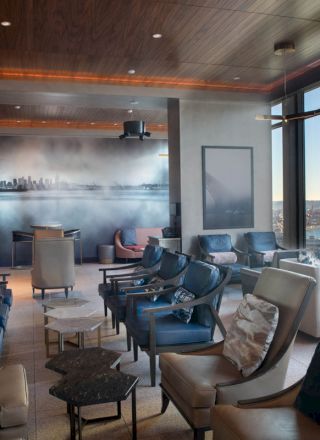 A stylish lounge with large windows showcasing a waterfront view, modern seating, and a mural on the wall. Natural light floods the space.