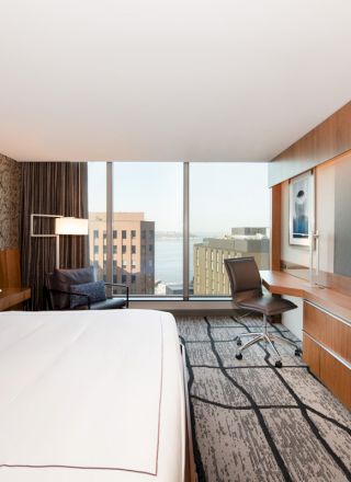 A modern hotel room with a large bed, desk, chair, and a wall-mounted TV, featuring large windows offering a city view.
