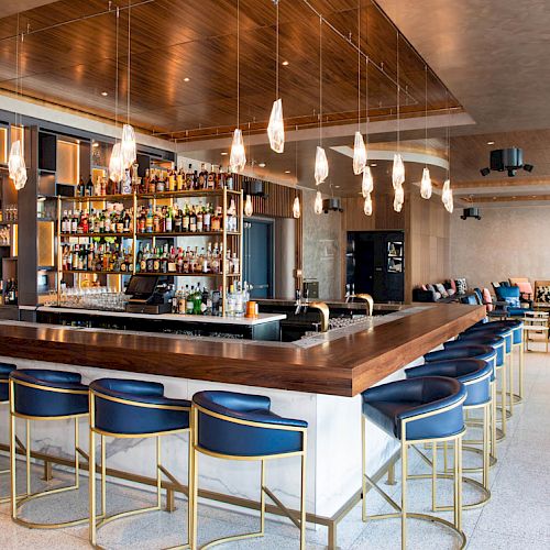 The image shows a modern bar with a wooden counter, blue and gold barstools, a well-stocked back bar, and hanging pendant lights, within a stylish lounge.