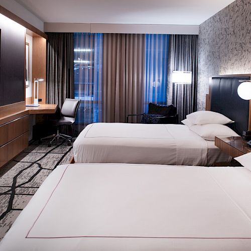 A modern hotel room with two beds, a TV, a desk, and lamps. The room features contemporary decor with a patterned carpet and wall design.