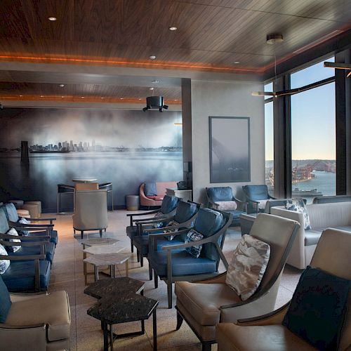 A modern lounge with large windows, ample seating, and a scenic view of a waterfront and ferris wheel outside the window, during the daytime.