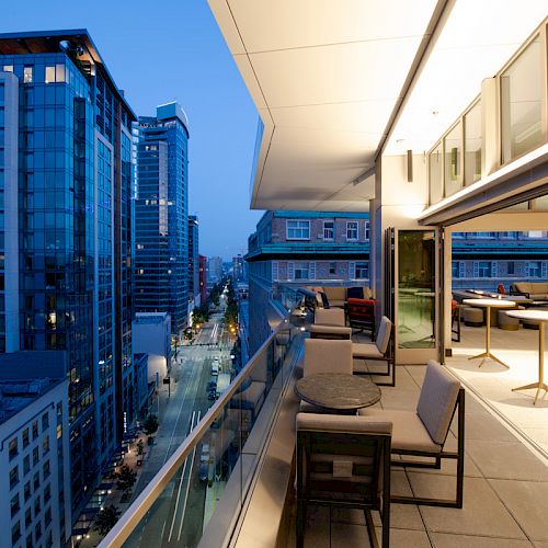 This image shows a modern rooftop terrace at twilight, featuring outdoor seating and tables, overlooking a city street with high-rise buildings.