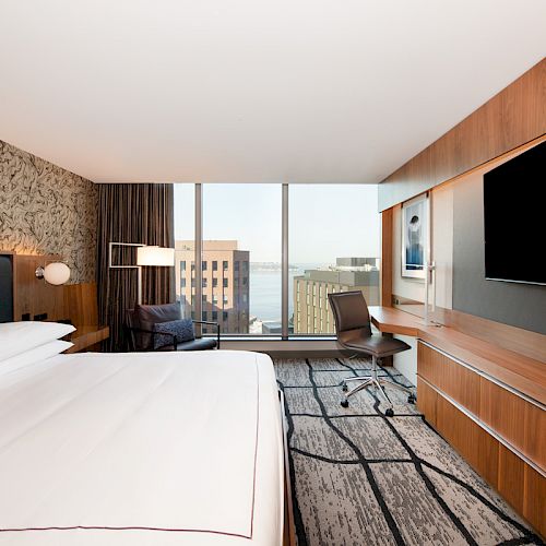 This image shows a modern hotel room with a king-sized bed, a desk with a chair, a large TV, and floor-to-ceiling windows with a city view.