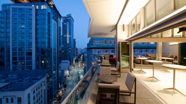 A modern balcony with seating and tables overlooking a cityscape with tall buildings at dusk, featuring floor-to-ceiling windows.