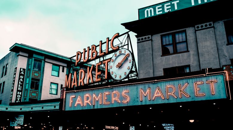 The image shows the iconic neon signs of the Public Market and Farmers Market, with a clock, located at Pike Place Market in Seattle, Washington.