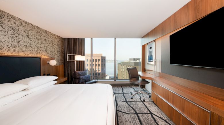 A modern hotel room with a large bed, wall-mounted TV, desk, chair, and floor-to-ceiling window offering a city view, featuring sleek decor.