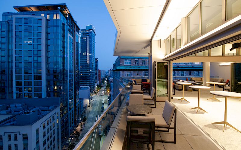 A modern balcony view of an urban cityscape at dusk, featuring glass buildings, outdoor seating, and tables under warm lighting.
