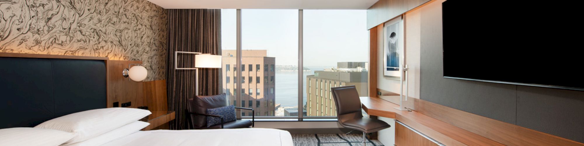 This image shows a modern hotel room with a large bed, a work desk, a big TV, and a view of city buildings through a window.