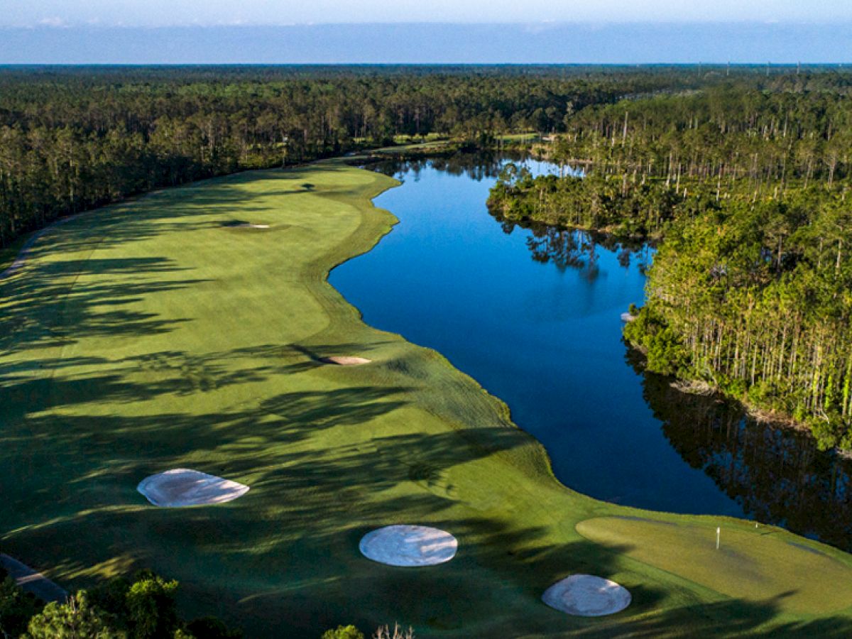 Aerial view of a golf course with sand bunkers, a fairway, and a water hazard surrounded by lush trees and a serene lake.