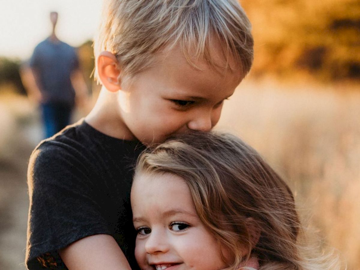 A young boy is hugging and kissing a younger girl on the head, both smiling, with a blurred adult figure in the background and a natural outdoor setting.